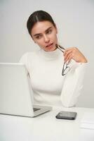 binnen- schot van jong mooi brunette zakenvrouw op zoek Bij scherm van haar laptop met verbaasd gezicht en Holding eyewear in verheven hand, poseren over- wit achtergrond foto