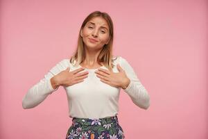 binnen- schot van positief lief blond dame in wit blouse en gebloeid rok op zoek Bij camera vriendelijke groet en Holding verheven palmen Aan haar borst, geïsoleerd over- roze achtergrond foto