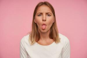 portret van mooi blond vrouw met gewoontjes kapsel fronsen wenkbrauwen en tonen haar tong terwijl poseren over- roze achtergrond, vervelend wit blouse foto