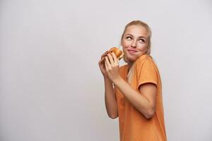 studio schot van hongerig verheugd jong blond vrouw in oranje t-shirt op zoek positief terzijde met mond vol van voedsel, Holding vers hamburger terwijl staand over- wit achtergrond foto