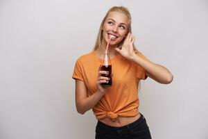 binnen- schot van blij mooi jong lang haren blond vrouw drinken Frisdrank en op zoek gelukkig terzijde met breed glimlach, wezen in mooi hoor humeur terwijl poseren tegen wit achtergrond foto