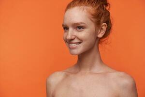 studio portret van jong aantrekkelijk leeskop vrouw met bun kapsel geïsoleerd over- oranje achtergrond, op zoek terzijde met charmant glimlach, demonstreren haar wit perfect tanden foto