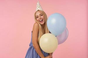 verheugd jong blond vrouw verheugt terwijl poseren in veelkleurig lucht ballonnen, vervelend feestelijk kleren en ijshoorntje hoed, geïsoleerd over- roze achtergrond, tonen haar aangenaam emoties foto