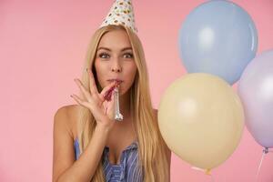 jong verheugd vrouw met lang blond haar- vieren verjaardag met veelkleurig lucht ballonnen, blazen partij toeter en glimlachen vriendelijke groet naar camera. mensen, vermaak en vakantie attributen foto