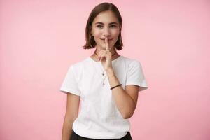 blij jong lief kort haren brunette vrouw in gewoontjes slijtage houden wijsvinger Aan haar lippen terwijl vragen naar houden haar geheim, staand over- roze achtergrond foto