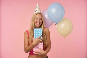 binnen- schot van positief jong lang haren vrouw verheugt terwijl poseren in veelkleurig lucht ballonnen, hebben pret Aan verjaardag partij en Holding geschenken, staand over- roze achtergrond foto