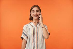 portret van een aantrekkelijk jong vrouw met glimlach aan het kijken Bij de camera. praten Aan een smartphone, vervelend gestreept shirt, tanden een beugel en armbanden. staand over- oranje achtergrond geïsoleerd foto