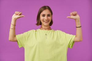 tiener- meisje, vrolijk en Vrolijk, met brunette kort haar. richten duimen Bij haarzelf en glimlach. staand over- Purper achtergrond. vervelend groen t-shirt, tanden een beugel, armbanden en ringen foto