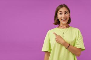 tiener- meisje, vrolijk en Vrolijk, met brunette kort haar, richten haar vinger naar de links Bij de kopiëren ruimte over- Purper achtergrond. tonen een teken. vervelend groen t-shirt, armbanden en ringen foto