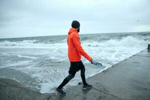 terug visie van jong gebaard Mens in zwart sportief kleren en warm oranje jas met kap wandelen langs kust Aan stormachtig grijs dag, houden geschiktheid fles met water in zijn hand- foto