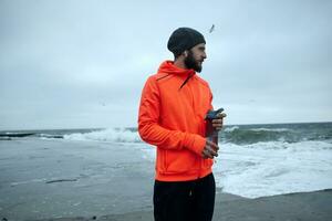 foto van jong actief brunette Mens met baard Holding fles met water in handen terwijl op zoek bedachtzaam Aan stormachtig zee, beginnend dag van ochtend- joggen voordat zijn werk