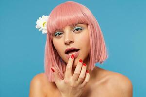 studio foto van jong lief roze haren dame met gekleurde bedenken Holding inhoudsopgave vinger Aan haar onderlip terwijl op zoek Bij camera met kalmte gezicht, staand tegen blauw achtergrond