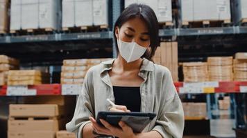 jonge azië zakenvrouw manager draagt gezichtsmasker op zoek naar goederen met behulp van digitale tablet controleren voorraadniveaus staan in retail winkelcentrum. distributie, logistiek, pakketten klaar voor verzending. foto