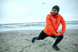 jong knap gebaard mannetje gekleed in sportief kleren en warm oranje jas poseren over- kustlijn van zee terwijl aan het doen uitrekken oefening, voorbereidingen treffen voor ochtend- training foto