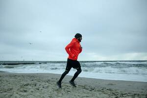 buitenshuis schot van jong gebaard loper met hoofdtelefoons jogging Aan zanderig strand voordat werken dag, Leidt gezond actief levensstijl, wezen kalmte en geconcentreerd foto