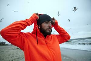 portret van aantrekkelijk jong donker haren Mens met baard gekleed in warm sportief kleren zetten Aan kap terwijl poseren over- strand Aan grijs bewolkt het weer, op zoek bedachtzaam verder foto