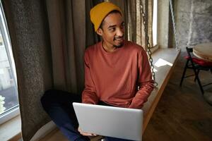 portret van positief knap donker huid mannetje met baard op zoek terzijde met licht glimlach, zittend Aan vensterbank met laptop, vervelend roze trui, blauw broek en mosterd pet foto