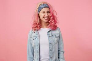 gelukkig schattig glimlachen dame met roze haar- en getatoeëerd handen, op zoek Bij de caera en knipogen, staand over- roze achtergrond, vervelend een wit t-shirt en denim jasje. foto