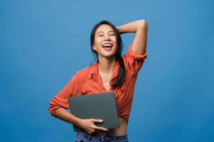 verraste jonge aziatische dame houdt laptop vast met positieve uitdrukking, glimlach breed, gekleed in casual kleding en kijkt naar camera op blauwe achtergrond. gelukkige schattige blije vrouw verheugt zich over succes. foto