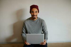 binnen- foto van mooi bruin ogen jong gebaard mannetje met donker huid houden laptop Aan zijn handen terwijl poseren over- wit muur, tonen zijn aangenaam emoties Aan camera