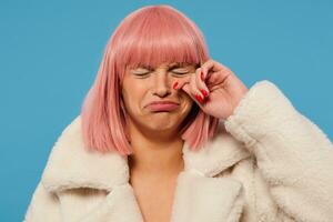 van streek jong aantrekkelijk vrouw met kort roze haar- draaien haar mond helaas terwijl huilen, houden haar ogen Gesloten terwijl afvegen tranen weg van haar wang, poseren over- blauw achtergrond in luxe kleren foto