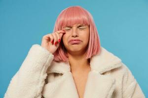 ongelukkig jong mooi roze haren vrouw met bob kapsel fronsen haar gezicht terwijl gaan naar schreeuw, houden ogen Gesloten en rozijn hand- naar haar gezicht, staand tegen blauw achtergrond foto