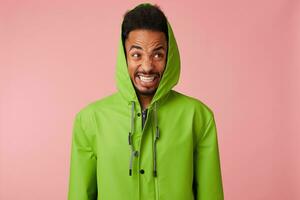 dichtbij omhoog van niet tevreden boos jong knap Afrikaanse Amerikaans knap vent in groen regen jas , staat over- roze achtergrond, barst zijn tanden agressief en looks in de camera, looks gek. foto