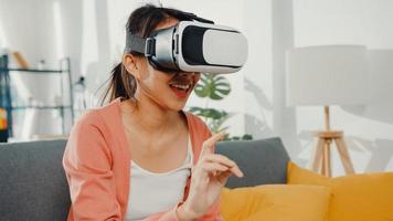 aziatische dame met een headsetbril van virtual reality gebarende hand zittend op de bank in de woonkamer in huis. blijf thuis covid quarantaine, re-imagining reality, vr-technologie van toekomstig concept. foto