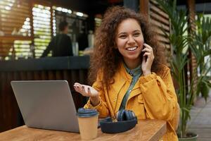 foto van jong donker huid gekruld vrouw locatie Aan een cafe terras, vervelend in geel jas, werken Bij een laptop, drinken koffie, glimlachen en pratend Aan de telefoon met een vriend.