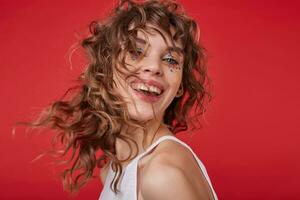 studio foto van charmant vrolijk jong vrouw met gekruld haar- draaien haar hoofd, vervelend wit top over- rood achtergrond, wezen in hoog geest en glimlachen blij