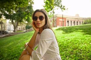 buitenshuis schot van charmant jong lang haren brunette vrouw in wit stip kleren op zoek Bij camera met licht glimlach, leunend haar hoofd Aan verheven hand- foto