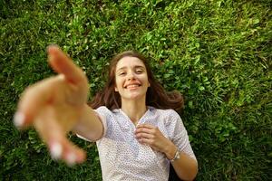 buitenshuis schot van lief jong brunette bruine ogen vrouw verhogen hand- naar camera en glimlachen op grote schaal, wezen in hoog geest terwijl poseren buitenshuis Aan zonnig warm dag foto