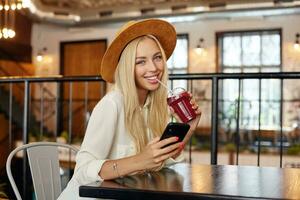 modieus mooi jong lang haren blond vrouw met mooi blauw ogen op zoek Bij camera met charmant glimlach, drinken smoothie met rietje terwijl zittend over- restaurant interieur foto