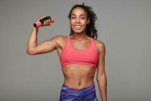 vrolijk jong lief donker huid brunette sportvrouw in atletisch slijtage op zoek gelukkig Bij camera en verhogen hand- met halter terwijl poseren over- grijs achtergrond foto