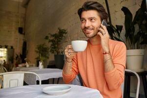 positief jong mooi mannetje in trui zittend in cafe met kop van thee, op zoek verder en glimlachen gelukkig, hebben aangenaam praten Aan mobiel telefoon foto