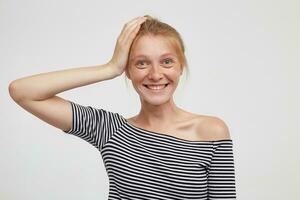 positief jong mooi roodharige vrouw met natuurlijk bedenken houden verheven palm Aan haar hoofd en op zoek Bij camera met charmant glimlach, staand tegen wit achtergrond foto