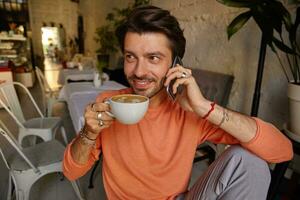 gelukkig aantrekkelijk mannetje met baard drinken koffie terwijl hebben gesprek Aan mobiel telefoon, poseren over- cafe interieur, wezen in mooi hoor humeur foto