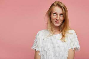 studio schot van verlegen jong aantrekkelijk vrouw met foxy haar- glimlachen aangenaam terwijl op zoek positief terzijde, vervelend wit elegant t-shirt terwijl poseren over- roze achtergrond foto