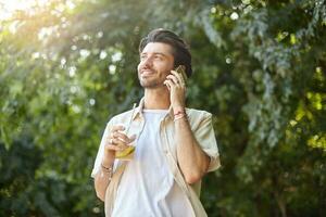 positief jong mooi Mens met baard poseren over- groen stad park Aan warm zonnig dag, maken telefoontje met zijn smartphone en Holding plastic kop met limonade foto