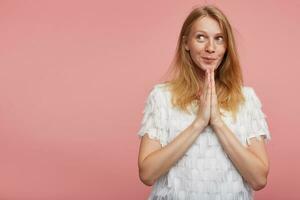 portret van jong mooi vrolijk vrouw met foxy haar- vouwen haar verheven palmen samen en op zoek positief terzijde met aangenaam glimlach, geïsoleerd over- roze achtergrond foto