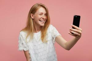 gelukkig jong lief roodharige vrouw met gewoontjes kapsel op zoek vrolijk Bij camera van haar smartphone en glimlachen breed terwijl maken selfie, staand tegen roze achtergrond foto