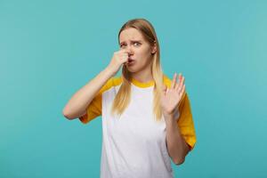 studio foto van jong blauwe ogen blond haar dame fronsen wenkbrauwen en sluitend haar neus- terwijl vermijden slecht geuren, geïsoleerd over- blauw achtergrond in gewoontjes t-shirt