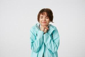 portret van positief jong mooi hoor dame in blauw regen jas, met gelukkig uitdrukkingen, met gesloten ogen en gebalde handen, hoopt voor geluk en dromen over mooi zo week, staat over- wit muur. foto