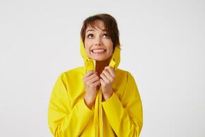 portret van jong gelukkig schattig kort haren meisje draagt in geel regen jas, schuilplaats onder een regen kap, breed glimlacht en looks omhoog, staat over- wit muur. foto
