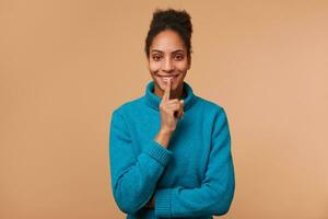 jong glimlachen Afrikaanse Amerikaans meisje met gekruld donker, demonstreert een gebaar van stilte, Holding een inhoudsopgave vinger in de buurt de mond oproepen voor privacy naar worden gehouden, geheim, blijven stil, kalm. foto