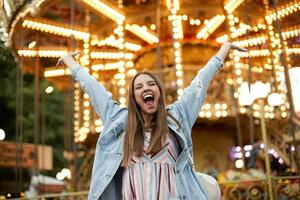 gelukkig mooi jong vrouw met lang bruin haar- poseren over- carrousel in park van attracties, verhogen handen gelukkig en glimlachen breed met Gesloten ogen, vervelend gewoontjes kleren foto