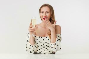 portret van jong vrolijk meisje met lang blond haar, met rood lippen in een stip jurk, verhogen een glas van Champagne, lacht en covers haar mond met hand- geïsoleerd over- wit achtergrond. foto