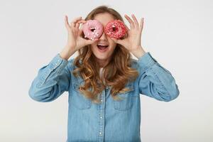 jong koel mannetje vervelend een denim shirt, met lang blond golvend haar, op zoek door donuts en glimlachen breed, geïsoleerd over- wit achtergrond. foto