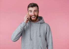 jong knap positief rood gebaard Mens in grijs capuchon, looks gelukkig en breed glimlacht, pratend Aan de telefoon met zijn vriendin, staat over- roze achtergrond. foto
