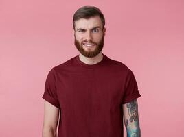 portret van jong knap fronsen misverstand rood gebaard Mens in rood t-shirt, staat over- roze achtergrond looks Bij de camera met walging. foto